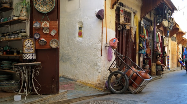 A narrow alley with shops displaying various traditional crafts and items. On the left, decorative plates and pottery are arranged on shelves and hanging on a door. Opposite the door, a small wooden cart with clay pots rests against a wall. The alley is lined with additional shops, displaying colorful fabrics and more handicrafts.