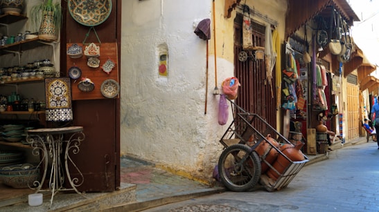 A narrow alley with shops displaying various traditional crafts and items. On the left, decorative plates and pottery are arranged on shelves and hanging on a door. Opposite the door, a small wooden cart with clay pots rests against a wall. The alley is lined with additional shops, displaying colorful fabrics and more handicrafts.