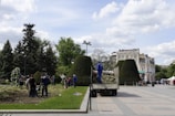 A group of people is working in a park-like setting, engaged in gardening activities. The scene features well-maintained hedges and trees, with some workers dressed in blue uniforms. In the background, there are several buildings with classic architecture under a partly cloudy sky.