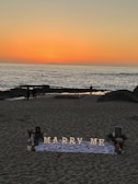 A romantic beach setting with a sunset backdrop over the ocean. Illuminated letters spelling 'Marry Me' are arranged on a white cloth adorned with rose petals. The scene includes a few people walking along the shoreline, and there are decorative elements like flowers and small lanterns around the setup.