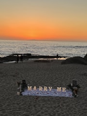 A romantic beach setting with a sunset backdrop over the ocean. Illuminated letters spelling 'Marry Me' are arranged on a white cloth adorned with rose petals. The scene includes a few people walking along the shoreline, and there are decorative elements like flowers and small lanterns around the setup.