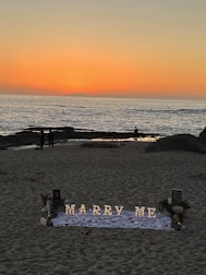 A romantic beach setting with a sunset backdrop over the ocean. Illuminated letters spelling 'Marry Me' are arranged on a white cloth adorned with rose petals. The scene includes a few people walking along the shoreline, and there are decorative elements like flowers and small lanterns around the setup.