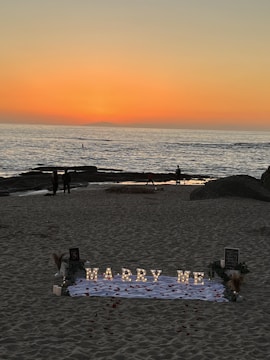 A romantic beach setting with a sunset backdrop over the ocean. Illuminated letters spelling 'Marry Me' are arranged on a white cloth adorned with rose petals. The scene includes a few people walking along the shoreline, and there are decorative elements like flowers and small lanterns around the setup.