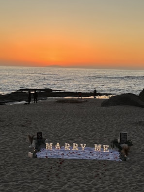 A romantic beach setting with a sunset backdrop over the ocean. Illuminated letters spelling 'Marry Me' are arranged on a white cloth adorned with rose petals. The scene includes a few people walking along the shoreline, and there are decorative elements like flowers and small lanterns around the setup.
