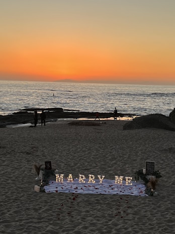 A romantic beach setting with a sunset backdrop over the ocean. Illuminated letters spelling 'Marry Me' are arranged on a white cloth adorned with rose petals. The scene includes a few people walking along the shoreline, and there are decorative elements like flowers and small lanterns around the setup.
