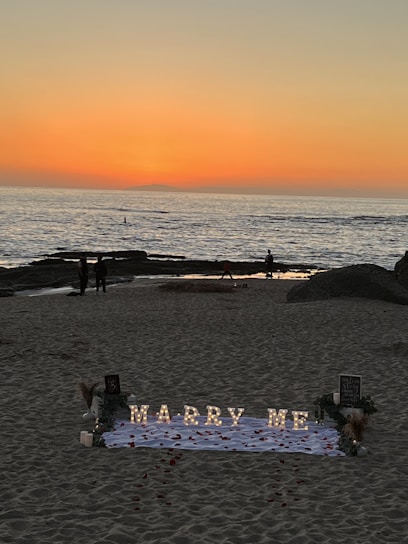 A romantic beach setting with a sunset backdrop over the ocean. Illuminated letters spelling 'Marry Me' are arranged on a white cloth adorned with rose petals. The scene includes a few people walking along the shoreline, and there are decorative elements like flowers and small lanterns around the setup.