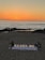 A romantic beach setting with a sunset backdrop over the ocean. Illuminated letters spelling 'Marry Me' are arranged on a white cloth adorned with rose petals. The scene includes a few people walking along the shoreline, and there are decorative elements like flowers and small lanterns around the setup.