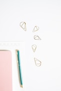 A minimalistic arrangement featuring a teal pen, pink notebook, golden geometric paper clips, and a white calendar or planner page. The objects are neatly placed on a light surface, giving a clean and organized appearance.
