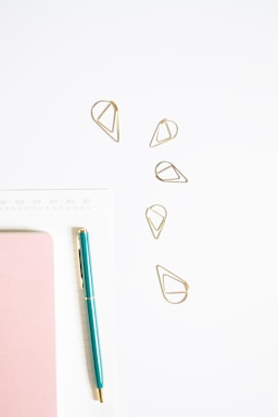 A minimalistic arrangement featuring a teal pen, pink notebook, golden geometric paper clips, and a white calendar or planner page. The objects are neatly placed on a light surface, giving a clean and organized appearance.