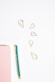 A minimalistic arrangement featuring a teal pen, pink notebook, golden geometric paper clips, and a white calendar or planner page. The objects are neatly placed on a light surface, giving a clean and organized appearance.