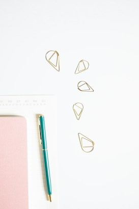 A minimalistic arrangement featuring a teal pen, pink notebook, golden geometric paper clips, and a white calendar or planner page. The objects are neatly placed on a light surface, giving a clean and organized appearance.
