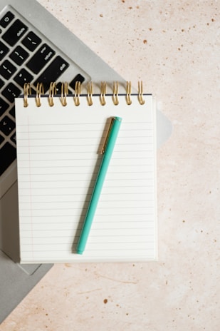 A spiral-bound notebook with lined pages is placed on top of a laptop keyboard. A turquoise pen lies diagonally across the notebook. The background is a light speckled surface.