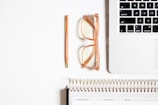 A neatly organized workspace featuring a section of a laptop keyboard, a pair of transparent glasses with peach-colored frames, a peach-colored pen with gold accents, and a spiral-bound planner with visible tabs for various sections.
