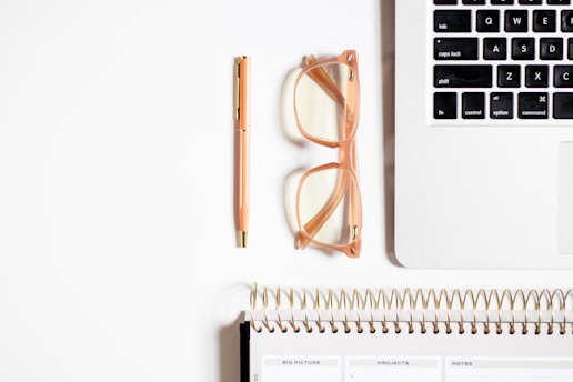 A neatly organized workspace featuring a section of a laptop keyboard, a pair of transparent glasses with peach-colored frames, a peach-colored pen with gold accents, and a spiral-bound planner with visible tabs for various sections.
