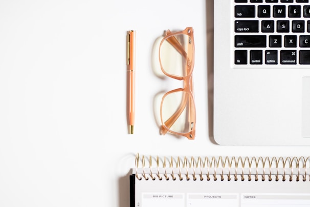 A neatly organized workspace featuring a section of a laptop keyboard, a pair of transparent glasses with peach-colored frames, a peach-colored pen with gold accents, and a spiral-bound planner with visible tabs for various sections.