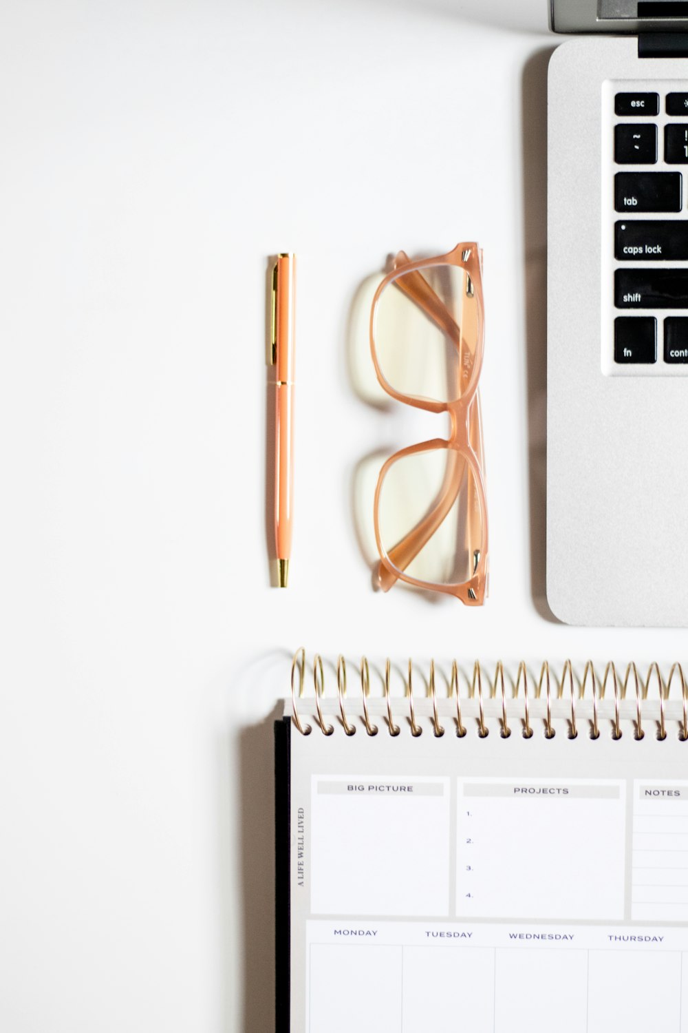 a desk with a notebook, glasses, pen and a laptop