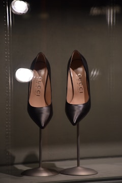 A pair of elegant black leather high-heeled shoes displayed on metal stands. The shoes appear sleek and sophisticated, with a glossy finish. Reflective lights above enhance their shine and highlight the brand name inside the shoes.