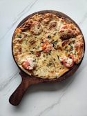 A freshly baked pizza topped with melted cheese, slices of tomato, and sprinkled herbs, set on a round, wooden pizza board with a handle. The board is placed on a smooth, white, marble-like surface.