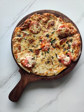 A freshly baked pizza topped with melted cheese, slices of tomato, and sprinkled herbs, set on a round, wooden pizza board with a handle. The board is placed on a smooth, white, marble-like surface.