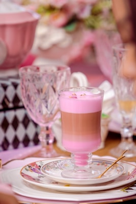 A layered dessert in a glass sits elegantly on a decorative plate. The dessert features a pink top layer with a brown layer beneath, suggesting a mix of flavors. The setting includes intricate glassware and delicate floral-patterned china, contributing to a sophisticated, pastel-themed ambiance.