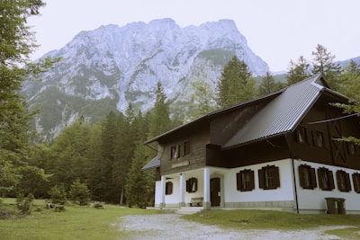 A rustic house with dark wooden and white exterior is situated at the edge of a lush green forest. The background features a towering mountain with rocky cliffs shrouded in light mist. The setting appears serene and secluded, amplifying the natural beauty and tranquility of the surroundings.