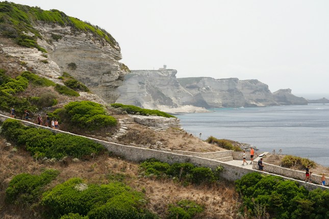 A coastal landscape with steep cliffs and lush greenery overlooking the sea. A stone pathway winds along the edge, where people are walking and admiring the view. The water is calm, and the sky is slightly overcast.