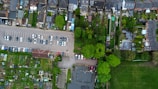 An aerial view depicts a residential neighborhood with rows of houses, a parking lot filled with cars, a variety of green spaces including gardens and a small park, alongside sections of open grass fields. The layout is organized with distinct areas for residential housing, parking, and green spaces.