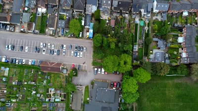 An aerial view depicts a residential neighborhood with rows of houses, a parking lot filled with cars, a variety of green spaces including gardens and a small park, alongside sections of open grass fields. The layout is organized with distinct areas for residential housing, parking, and green spaces.