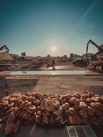 An outdoor market scene with various bread and baked goods arranged on trays at the foreground. In the background, there's a vast open area with a few vehicles, construction machinery, and two people standing near each other. The sun is low in the sky, casting long shadows, indicating either early morning or late afternoon. Buildings and other urban structures are visible in the distance.