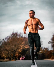 A fit man with a muscular build is running down a deserted road with autumn trees on either side. He is wearing black sports shorts and leggings. The sky is cloudy, creating a moody backdrop.