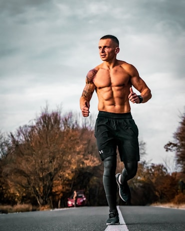A fit man with a muscular build is running down a deserted road with autumn trees on either side. He is wearing black sports shorts and leggings. The sky is cloudy, creating a moody backdrop.