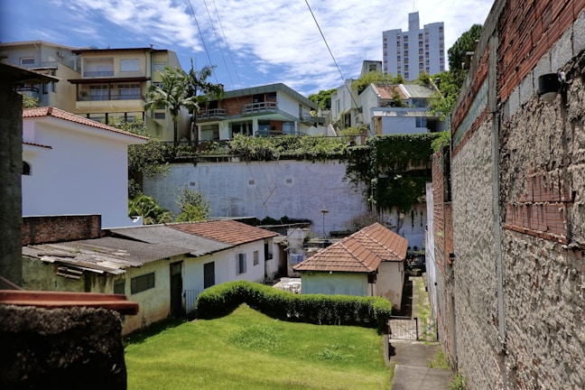 A residential area with a mix of different housing structures, including older brick houses with tiled roofs and more modern multifloor buildings in the background. A green lawn and some trees are visible, alongside brick and concrete walls. Overhead power lines stretch across the scene, and the sky is partially cloudy.