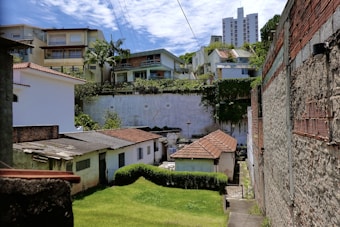 A residential area with a mix of different housing structures, including older brick houses with tiled roofs and more modern multifloor buildings in the background. A green lawn and some trees are visible, alongside brick and concrete walls. Overhead power lines stretch across the scene, and the sky is partially cloudy.