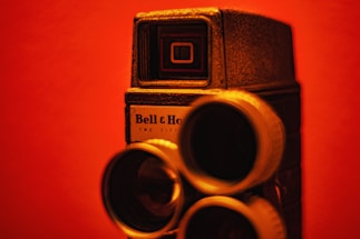 A vintage motion picture camera with a textured metallic body and multiple lenses is set against a red background. The camera displays the brand name 'Bell & Howell' and features intricate details that evoke a sense of classic technology.