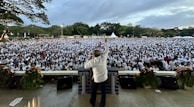 A large crowd gathers in an outdoor setting, with numerous people wearing white shirts. A person stands facing the crowd from a stage, raising an arm as if addressing the audience. The setting appears to include greenery and open skies, with some decorative plants and floral arrangements at the front of the stage.