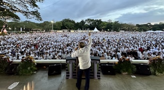 A large crowd gathers in an outdoor setting, with numerous people wearing white shirts. A person stands facing the crowd from a stage, raising an arm as if addressing the audience. The setting appears to include greenery and open skies, with some decorative plants and floral arrangements at the front of the stage.