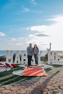 A couple stands on a decorated pathway by the sea, surrounded by large white letters spelling out 'MARRY ME'. The scene includes a red carpet lined with candles and rose petals, with a background of a clear blue sky and ocean.