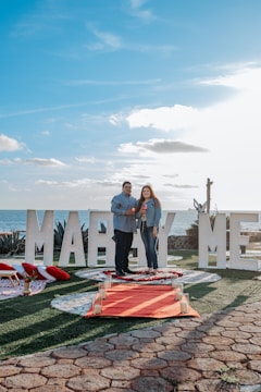 A couple stands on a decorated pathway by the sea, surrounded by large white letters spelling out 'MARRY ME'. The scene includes a red carpet lined with candles and rose petals, with a background of a clear blue sky and ocean.