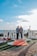 A couple stands on a decorated pathway by the sea, surrounded by large white letters spelling out 'MARRY ME'. The scene includes a red carpet lined with candles and rose petals, with a background of a clear blue sky and ocean.