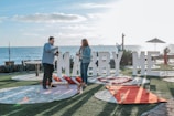 A couple stands near the ocean with a large sign reading 'Marry Me' in the background. They are on a grassy area with stone pathways, surrounded by candles and a red carpet leading up to the sign. The sky is clear, and the scene is set for a proposal, featuring a picnic setup with a blanket and pillows.