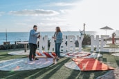 A couple stands near the ocean with a large sign reading 'Marry Me' in the background. They are on a grassy area with stone pathways, surrounded by candles and a red carpet leading up to the sign. The sky is clear, and the scene is set for a proposal, featuring a picnic setup with a blanket and pillows.