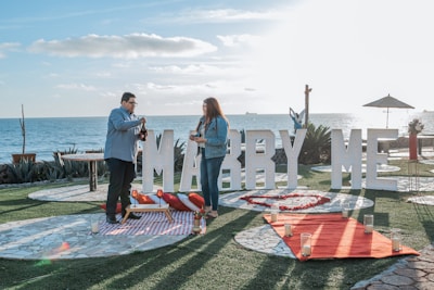 A couple stands near the ocean with a large sign reading 'Marry Me' in the background. They are on a grassy area with stone pathways, surrounded by candles and a red carpet leading up to the sign. The sky is clear, and the scene is set for a proposal, featuring a picnic setup with a blanket and pillows.
