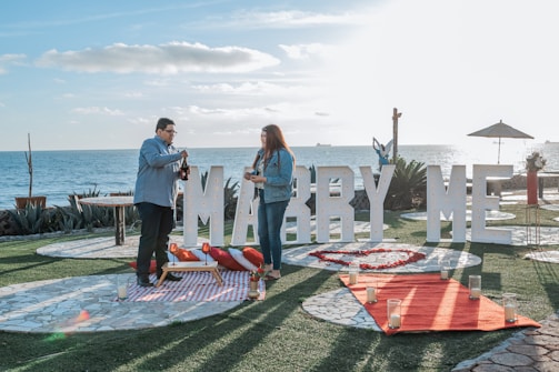 A couple stands near the ocean with a large sign reading 'Marry Me' in the background. They are on a grassy area with stone pathways, surrounded by candles and a red carpet leading up to the sign. The sky is clear, and the scene is set for a proposal, featuring a picnic setup with a blanket and pillows.