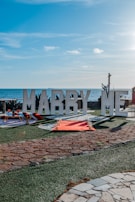 Large white letters spelling 'MARRY ME' are set up on a grassy outdoor area overlooking the sea. A red carpet leads up to the letters, with candles arranged along the sides. The sky is clear and blue, contributing to a romantic setting.