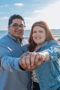 A couple is holding hands while smiling at the camera. The setting appears to be a beach with a clear blue sky and ocean in the background. Both individuals are dressed casually, and the woman's denim jacket complements her floral top.