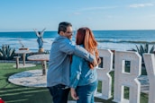 A couple embraces near a scenic oceanfront, with a view of the blue sea under a clear sky. Large white letters with lights are in the foreground on a green grassy area. A statue of an angel is visible in the background along with some outdoor tables and chairs surrounded by plants.