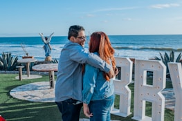 A couple embraces near a scenic oceanfront, with a view of the blue sea under a clear sky. Large white letters with lights are in the foreground on a green grassy area. A statue of an angel is visible in the background along with some outdoor tables and chairs surrounded by plants.