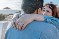 A close-up image of two people embracing. The woman's hand is prominently displayed, adorned with a ring, as she wraps her arms around the man's neck. Both are dressed in casual attire, with the woman gazing into the man's eyes affectionately.