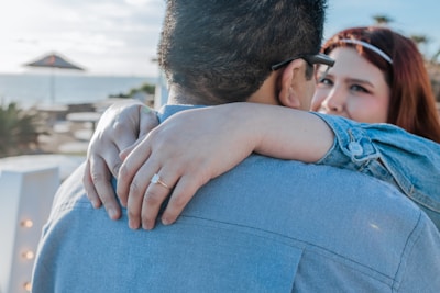A close-up image of two people embracing. The woman's hand is prominently displayed, adorned with a ring, as she wraps her arms around the man's neck. Both are dressed in casual attire, with the woman gazing into the man's eyes affectionately.
