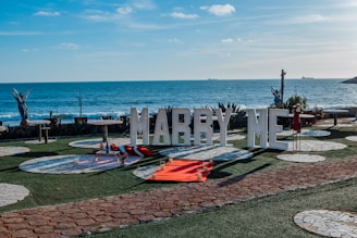 A romantic seaside setup features large letters spelling 'MARRY ME' in front of an ocean backdrop. Decorative elements include a picnic setting with a red blanket and cushions, surrounded by white circular stone patterns on the grass. There are also small candle holders placed around the blanket, adding to the intimate atmosphere. Two angel statues stand on either side, and there are ships visible in the distance on the ocean.